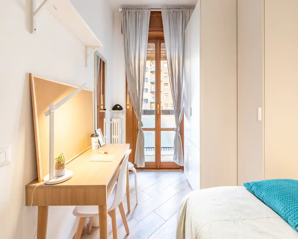 Camera singola luminosa e moderna con scrivania in legno vicino alla finestra, letto ben rifatto con coperta turchese e armadio a muro — ideale per uno studente o occupante singolo.