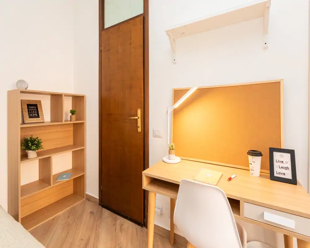Angolo di camera con scrivania in legno e scaffale; area di lavoro illuminata con lampada, pannello in sughero e piccole piante decorativi.