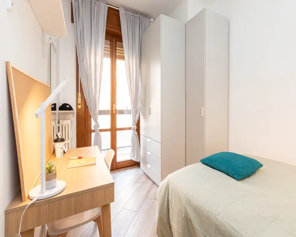 Camera singola luminosa e compatta con letto, armadio e scrivania vicino alla porta balcone. Luce naturale e arredi neutri rendono l'ambiente accogliente e funzionale, ideale per uno studente o una persona sola.