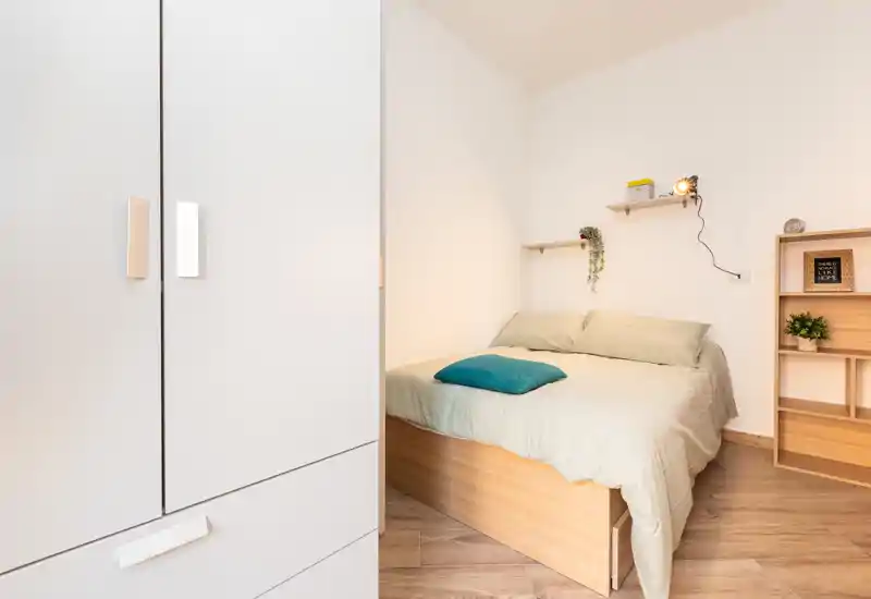 Camera da letto compatta e luminosa con letto matrimoniale su base in legno, biancheria neutra e cuscino turchese. Armadio bianco e mensole in legno chiaro per un aspetto minimal e ordinato.