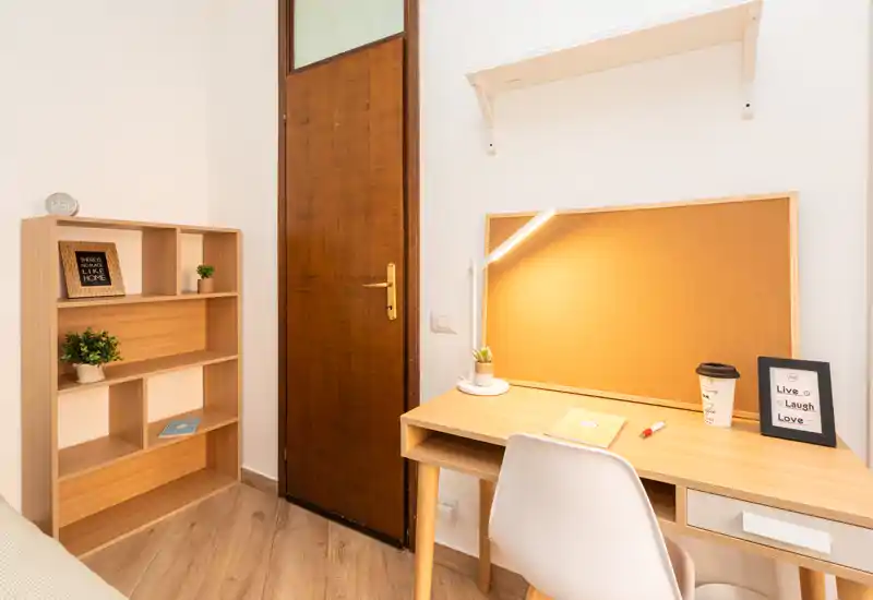 Angolo di camera con scrivania in legno e scaffale; area di lavoro illuminata con lampada, pannello in sughero e piccole piante decorativi.