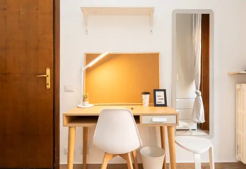 Angolo letto/scrivania ordinato in un luminoso angolo della camera, con una scrivania in legno, lampada, pannello in sughero e specchio a figura intera — ideale per studiare o prepararsi.
