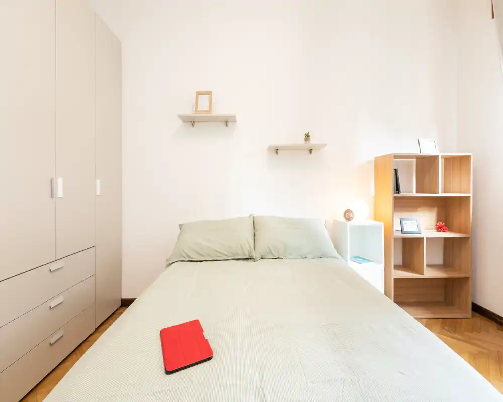 Dormitorio luminoso y minimalista con una gran cama doble en el centro, ropa de cama verde suave, armario a la izquierda y estantería abierta con lámpara a la derecha — ideal para anuncios de alquiler que destaquen la simplicidad y la luz natural.