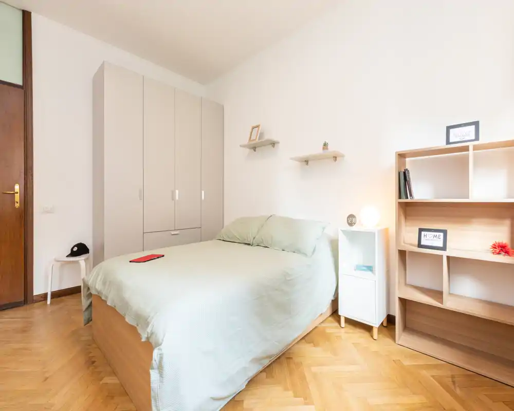 Dormitorio luminoso y ordenado con cama doble, ropa de cama clara y suelos de madera. La habitación incluye armario, mesita con lámpara y estanterías: un espacio tranquilo y bien iluminado, ideal para alquileres de corta estancia.