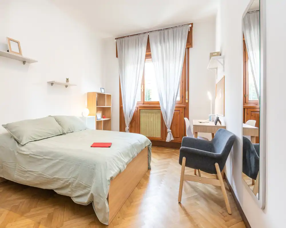 Dormitorio luminoso y acogedor con cama doble, suelos de madera, un sillón y un escritorio junto a la ventana. La luz natural y los tonos neutros crean un ambiente tranquilo perfecto para anuncios.