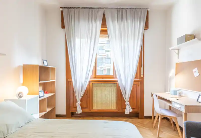 Camera da letto luminosa e curata con letto matrimoniale, cornice finestra in legno e un piccolo angolo scrivania — ideale per riposo e smart working.
