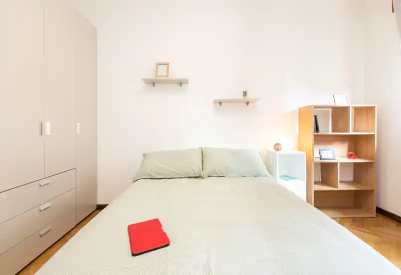 Camera da letto luminosa e minimal con un grande letto matrimoniale al centro, biancheria verde chiaro, armadio a sinistra e scaffalatura aperta con lampada a destra — ideale per annunci in affitto che valorizzano semplicità e luce naturale.