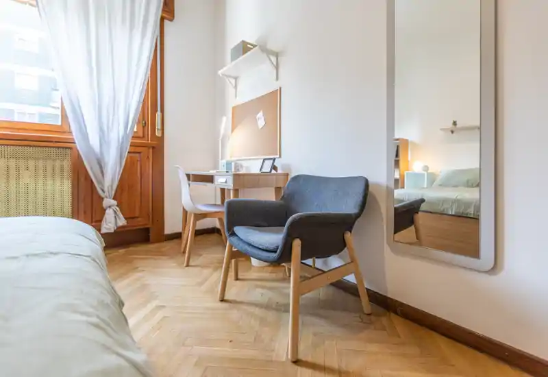 Angolo della camera luminoso e accogliente con letto comodo, poltrona per la lettura e scrivania vicino alla finestra che lascia entrare luce naturale — pavimenti in legno caldo e arredamento minimal creano un’atmosfera rilassante.
