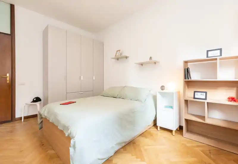 Camera da letto luminosa e ordinata con letto matrimoniale, biancheria chiara e pavimento in legno. L'ambiente offre un armadio, comodino con lampada e mensole: spazio tranquillo e ben illuminato ideale per affitti brevi.