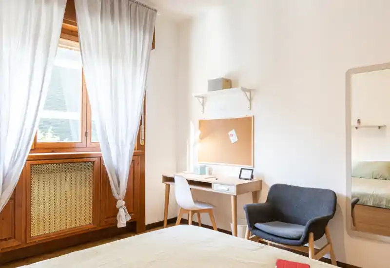Camera da letto luminosa e accogliente con luce naturale, letto rifatto, una piccola scrivania in legno e una comoda poltrona — perfetta per riposo e lavoro da remoto.