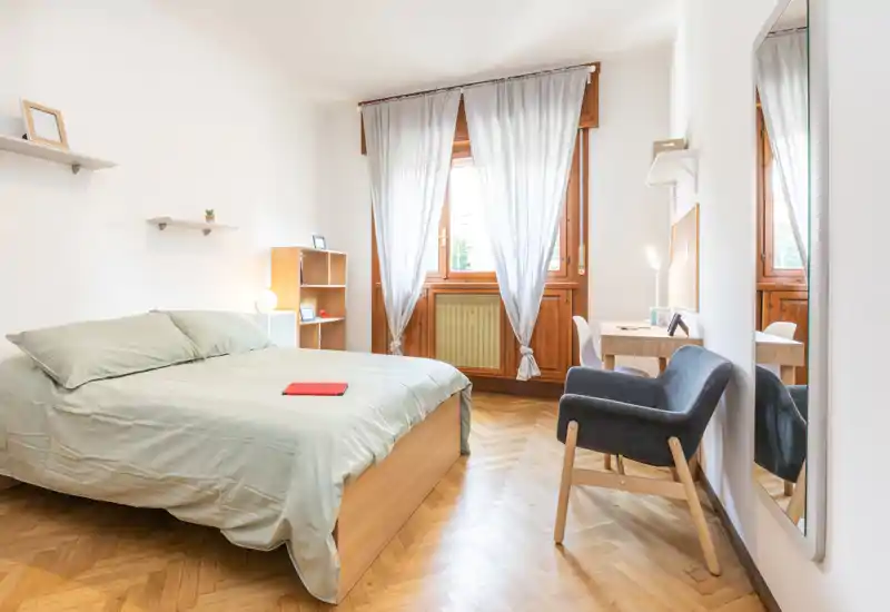 Camera da letto luminosa e accogliente con letto matrimoniale, pavimento in legno, poltrona per la lettura e scrivania vicino alla finestra. La luce naturale e i toni neutri creano un'atmosfera tranquilla perfetta per gli annunci.