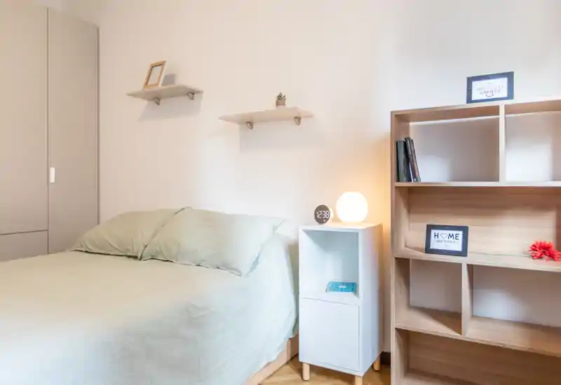 Camera da letto luminosa e ben arredata con letto completamente visibile, biancheria morbida, comodino e mensole—perfetta per un annuncio accogliente.