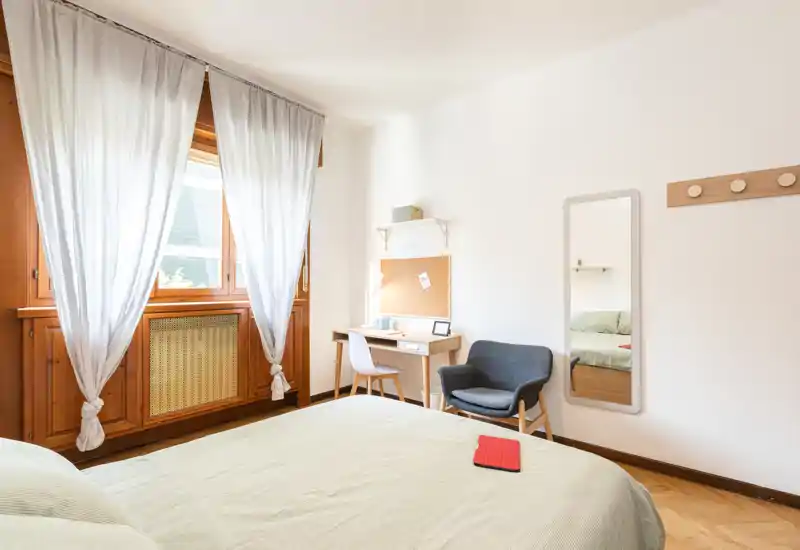 Camera da letto luminosa e minimal con letto ampio, infissi in legno, una piccola scrivania e una poltrona accogliente — perfetta per riposo e smart working.