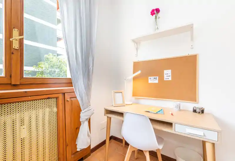 Angolo luminoso con finestra dal telaio in legno e una scrivania ordinata davanti a una bacheca in sughero — ideale come studio o postazione di lavoro.
