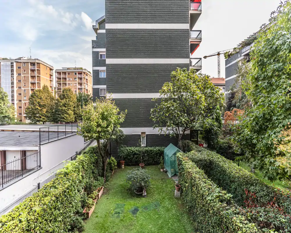 Jardín privado en planta baja con setos recortados, césped pequeño, macetas y un invernadero compacto—ideal para descansar o para jardinería ligera.