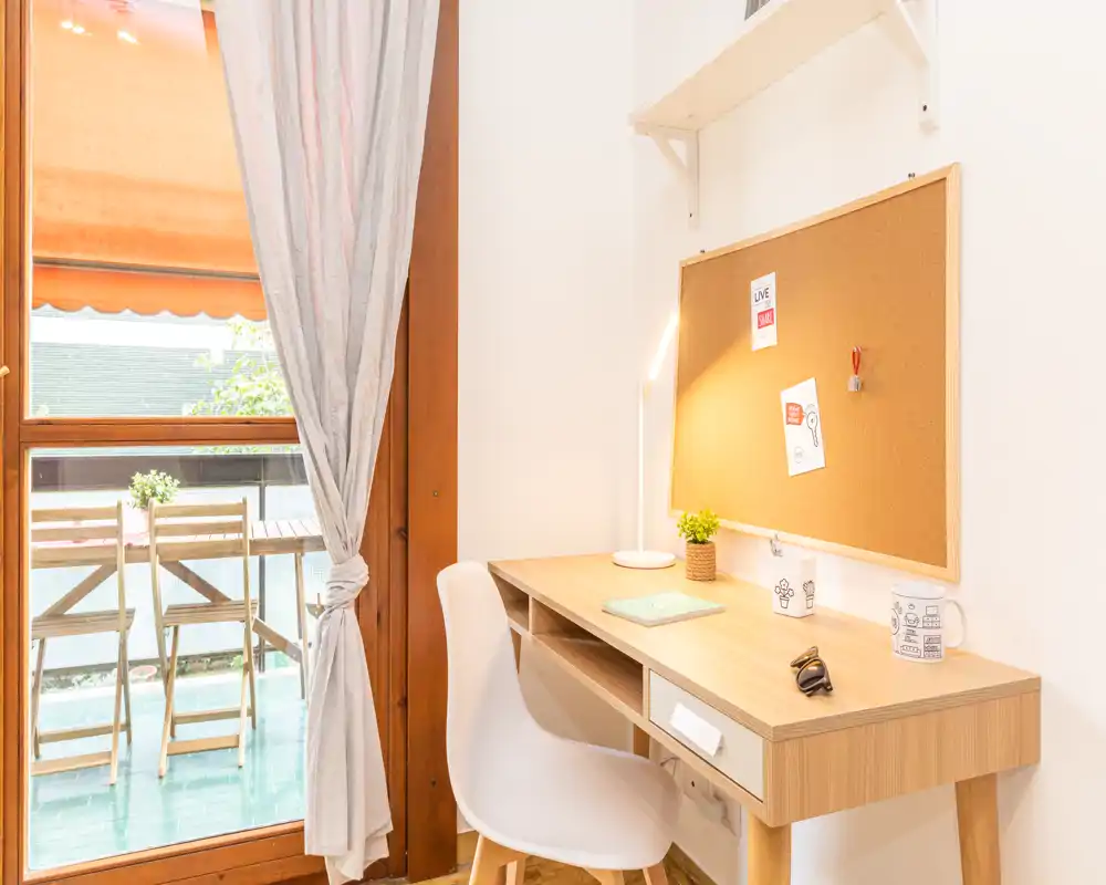 Rincón de trabajo luminoso y ordenado con escritorio de madera, panel de corcho y silla junto a una puerta de cristal que da a un pequeño balcón.