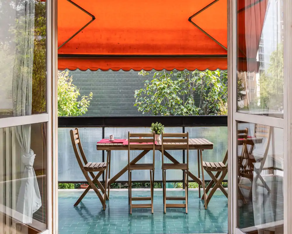 Balcón soleado con mesa de madera y sillas plegables bajo un toldo naranja brillante — ideal para desayunos o comidas informales al aire libre.