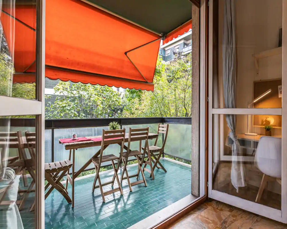 Balcón soleado con mesa de madera pequeña y sillas plegables, suelo de baldosa verde y toldo naranja retráctil — ideal para el café de la mañana.