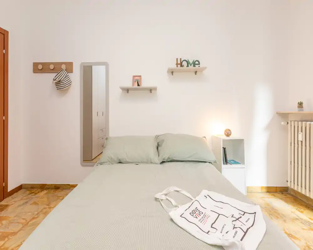 Dormitorio luminoso y ordenado con cama doble completa, ropa de cama suave, estantería junto a la cama e iluminación cálida — perfecto para una estancia acogedora.