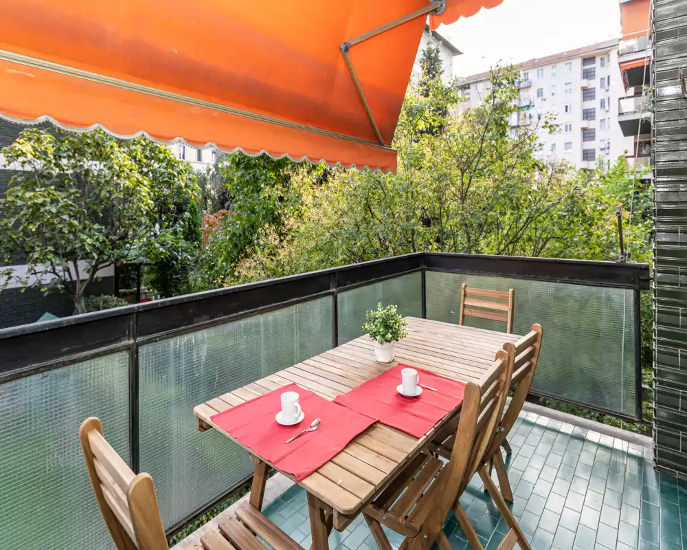 Balcón soleado con mesa de madera para cuatro, individuales rojos, una planta en maceta y un toldo naranja retráctil con vistas a árboles y edificios cercanos.
