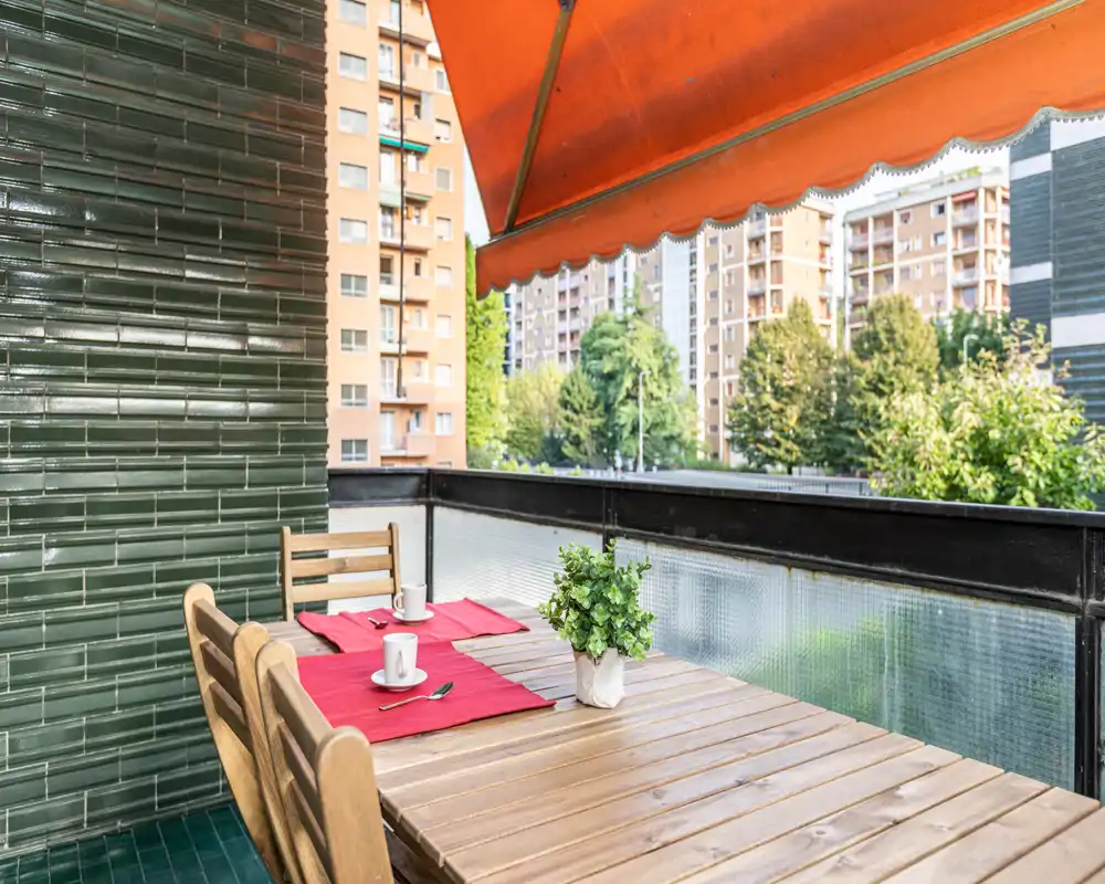 Balcón soleado amueblado con mesa de madera, dos sillas, manteles rojos y una pequeña planta en maceta; vistas a edificios residenciales y zonas verdes.