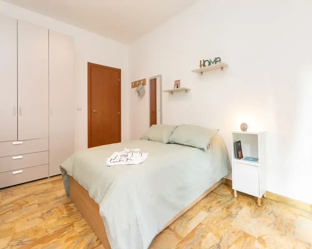 Dormitorio luminoso y ordenado con cama de matrimonio, ropa de cama verde suave y un mueble auxiliar sencillo; paredes neutras y armario que crean un ambiente moderno y tranquilo.