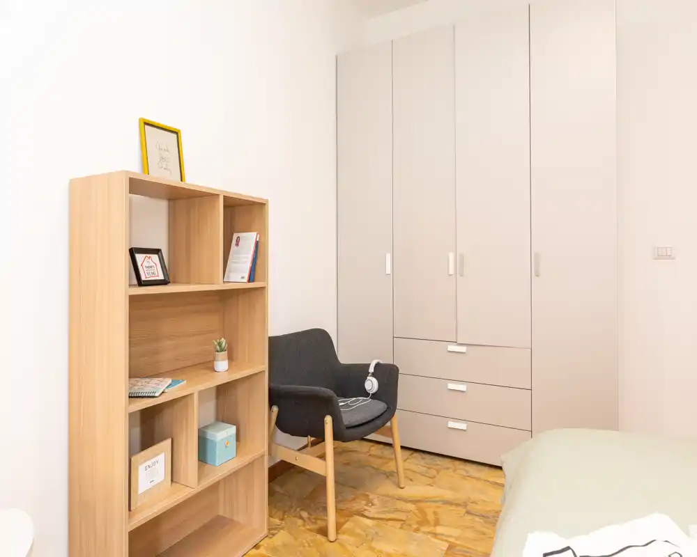Rincón de dormitorio luminoso y minimalista con cama visible, armario y un butacón acogedor. Estantería de madera y suelo cálido aportan un toque agradable.