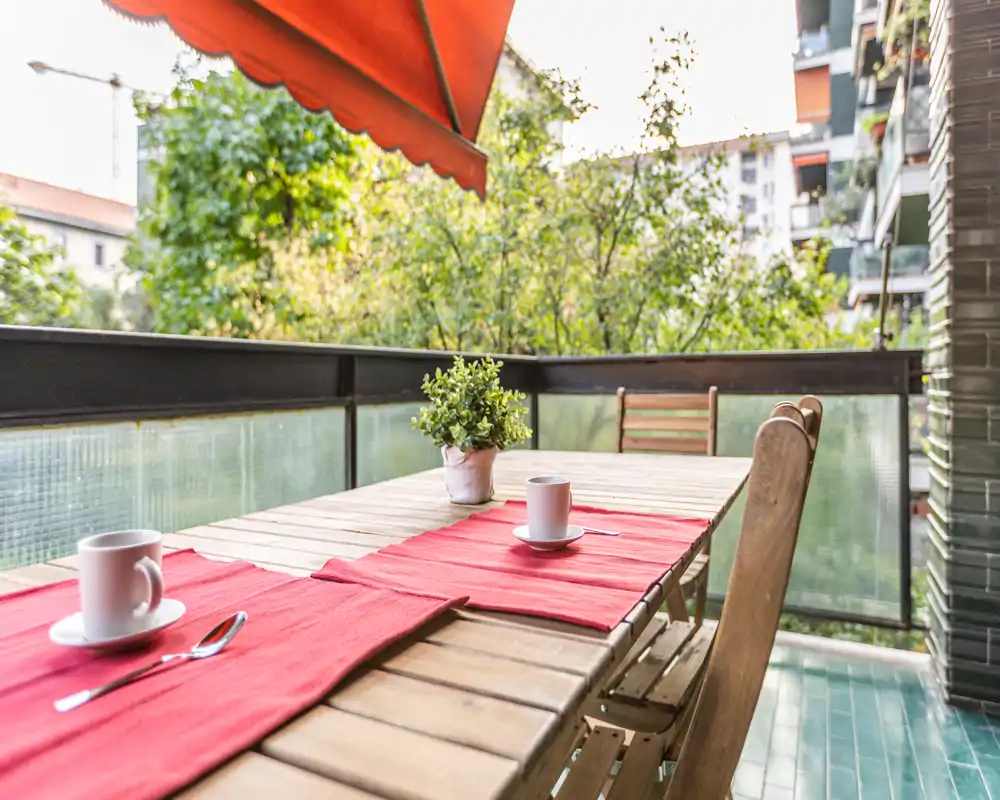 Balcón soleado con mesa de madera para dos, manteles rojos, planta en maceta y toldo naranja retráctil: perfecto para desayunos al aire libre.