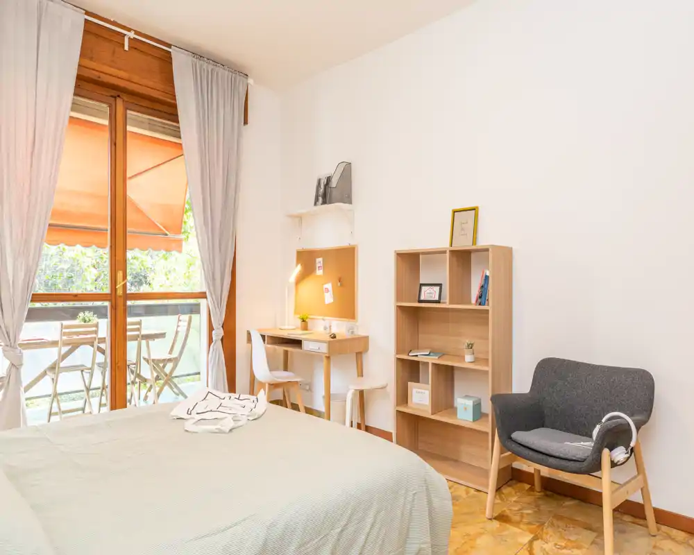 Dormitorio luminoso y acogedor con una cama grande en primer plano, un pequeño escritorio, estantería y un sillón cómodo. Grandes puertas correderas dan a un balcón que aporta abundante luz natural.