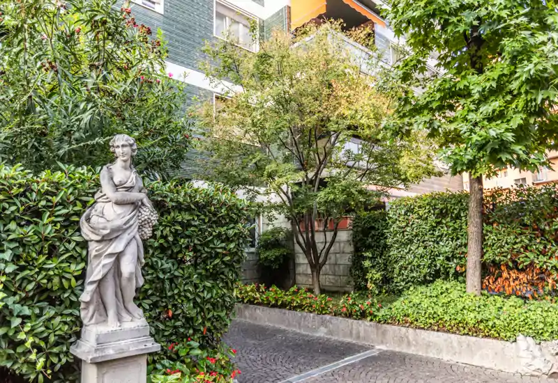 Cortile paesaggistico con alberi maturi, siepi fitte e una statua classica decorativa accanto a un vialetto lastricato — uno spazio esterno verde e tranquillo davanti a un edificio residenziale.