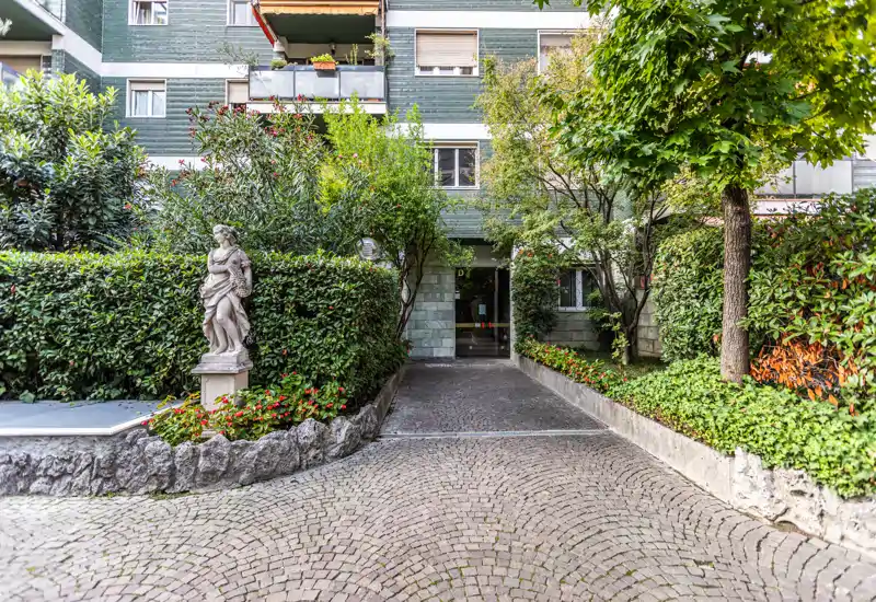Ingresso dell'edificio gradevole con vialetto lastricato, siepi curate, aiuole fiorite e una statua decorativa che conduce alla porta principale.
