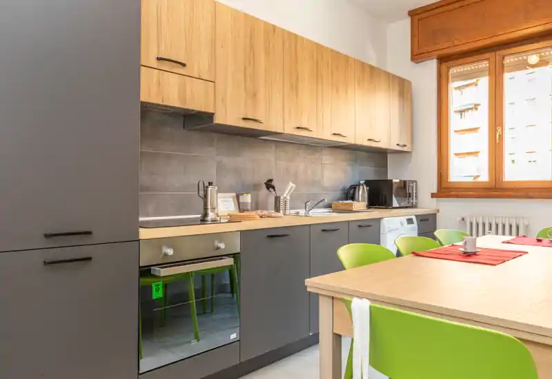 Cucina moderna con mobili in legno e grigio scuro, forno incassato e lavatrice, tavolo da pranzo con sedie verde brillante illuminato da luce naturale attraverso una grande finestra.