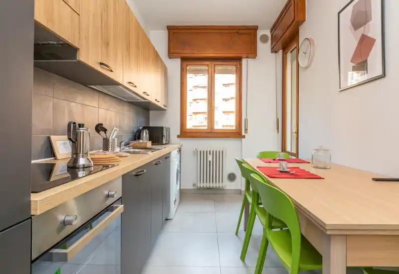 Cucina moderna e luminosa a corridor con pensili in legno, elettrodomestici integrati e un piccolo tavolo da pranzo con sedie colorate — perfetta per appartamenti compatti.