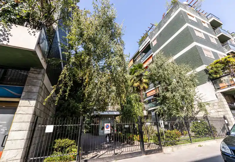 Ingresso condominiale recintato con alberi maturi e giardino curato; facciata di un edificio pluripiano visibile sotto luce diurna chiara.