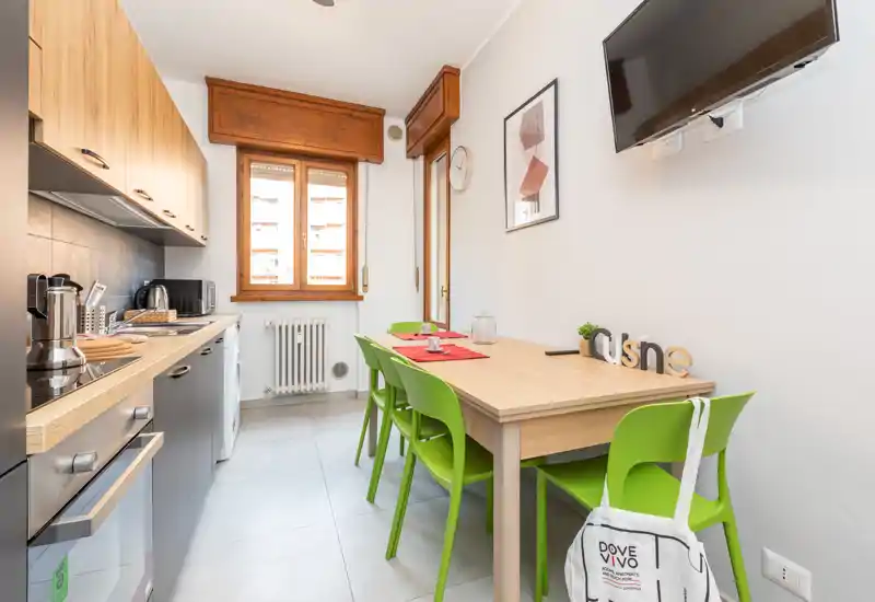 Cucina luminosa e moderna con piani in legno chiaro, elettrodomestici e quattro sedie verdi attorno al tavolo. La luce naturale dalla finestra valorizza l'area pranzo, perfetta per piccoli gruppi.