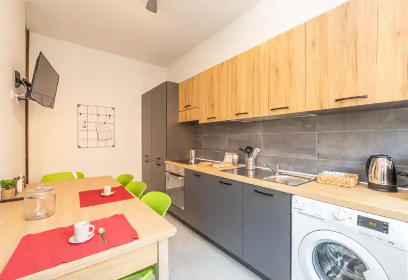 Cucina moderna attrezzata con pensili in legno, basi grigie e tavolo da pranzo con tovagliette rosse; ambiente luminoso e ordinato con elettrodomestici integrati e lavatrice.