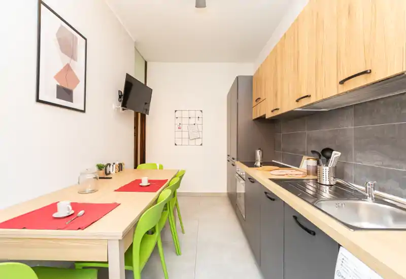 Cucina a corridoio moderna con lunga superficie di lavoro, lavello integrato e elettrodomestici, tavolo da pranzo e sedie verdi — ordinata, luminosa e pronta all'uso.