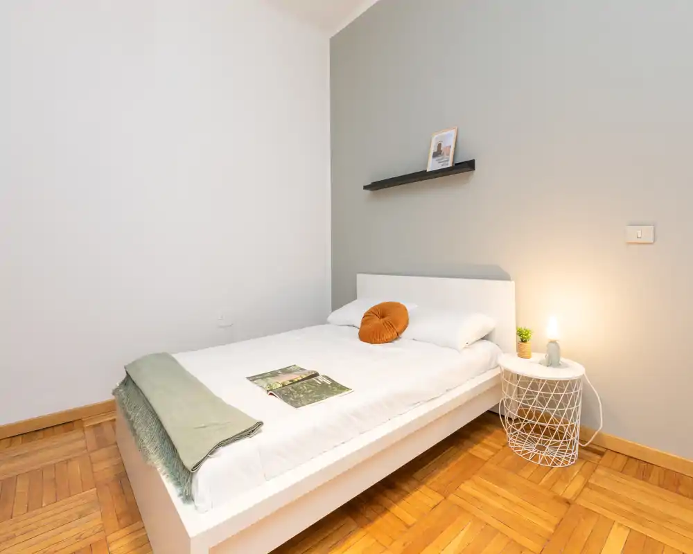 Camera da letto singola luminosa e minimale con letto bianco su piattaforma, pavimento in parquet caldo e illuminazione soffusa al comodino. Arredata con un cuscino rotondo, una piccola pianta e una mensola con cornice, risulta tranquilla e accogliente.