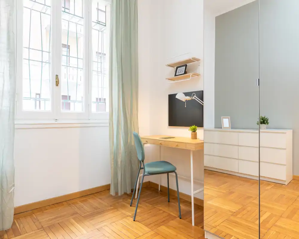 Angolo camera luminoso e minimal con una scrivania in legno, sedia, cassettiera e grande armadio con anta specchiata che riflette il parquet e la luce della finestra.