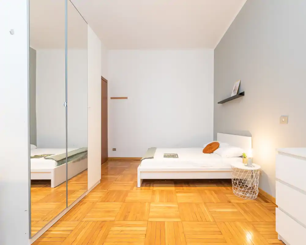 Camera da letto minimalista e luminosa con letto basso bianco, pavimento in parquet e armadio con ante a specchio che amplificano la profondità.
