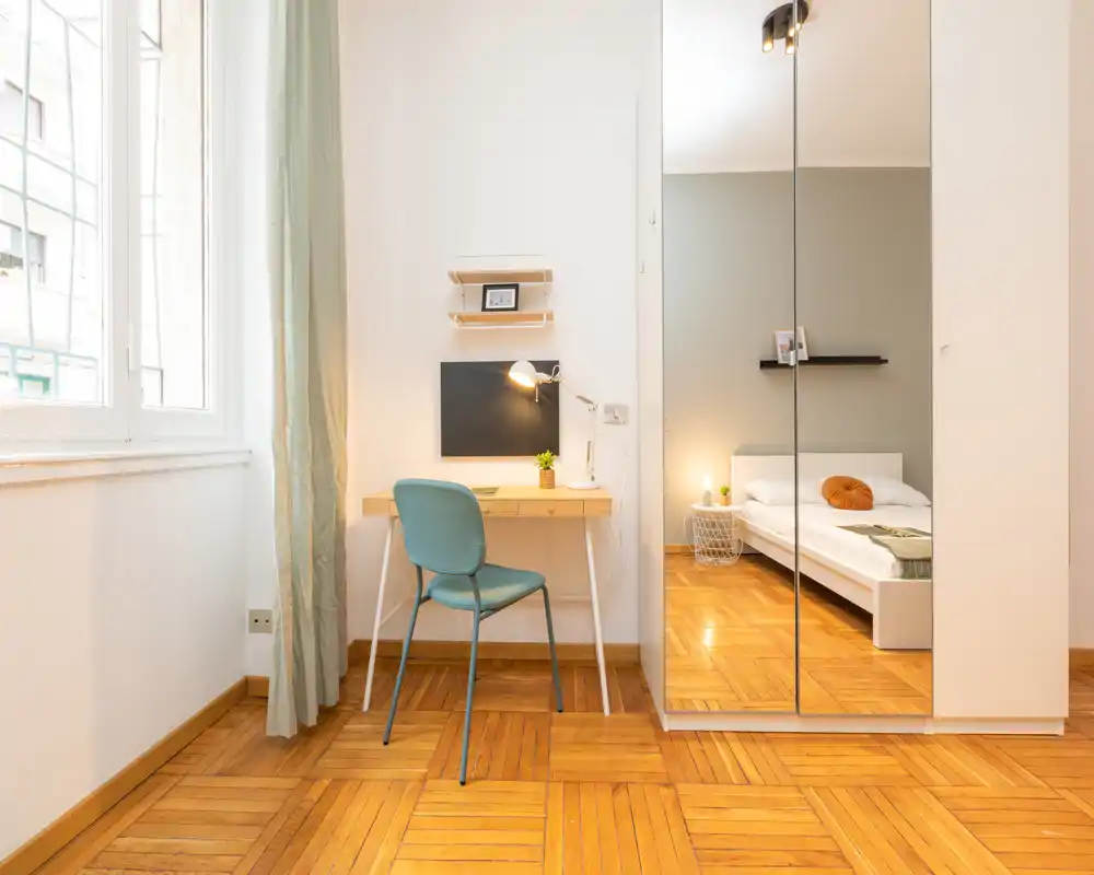 Camera da letto moderna e luminosa con un letto comodo, lampada da comodino e armadio con ante a specchio che ampliano lo spazio; piccolo scrittoio vicino alla finestra.
