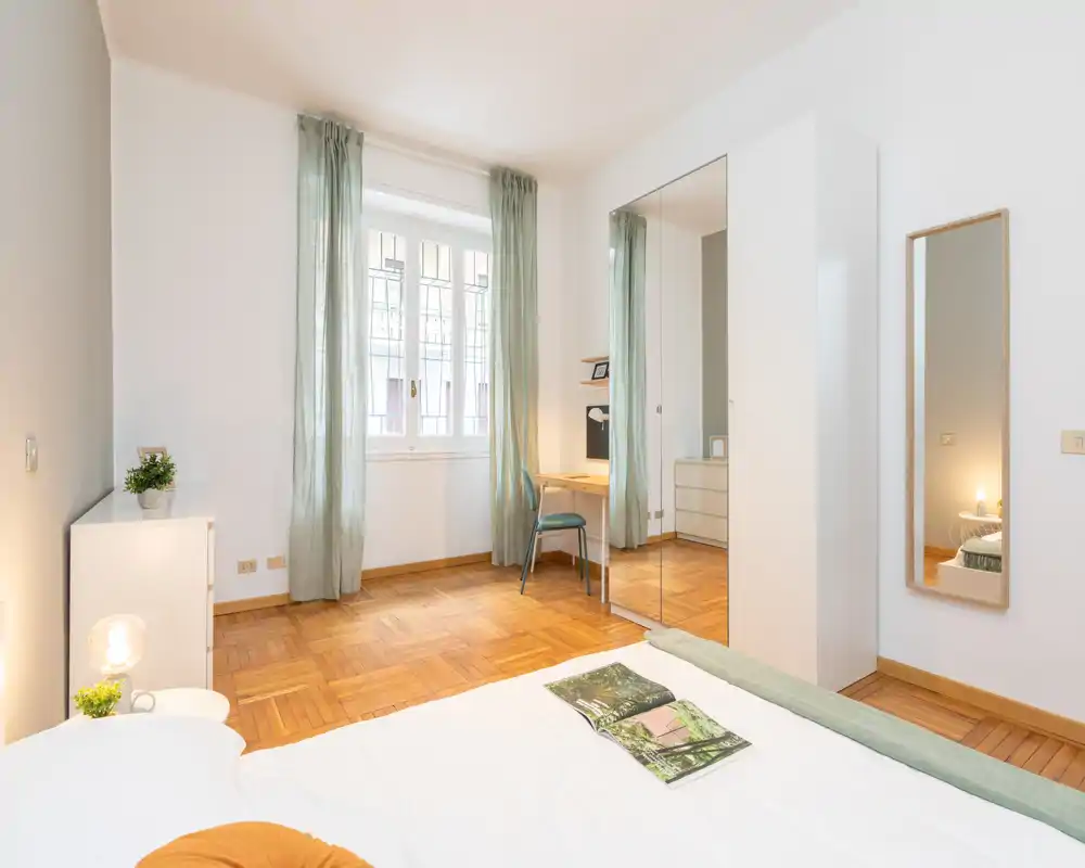 Camera da letto luminosa e moderna con un comodo letto matrimoniale, pavimento in parquet, ampia finestra con tende leggere e armadio con specchio che dona profondità.