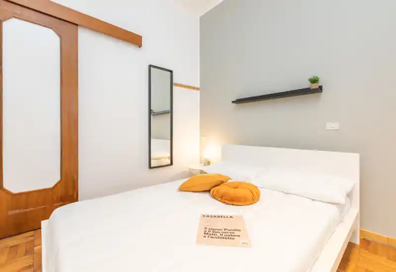 Dormitorio moderno y luminoso con cama doble blanca, ropa de cama limpia, cojines mostaza y una pequeña estantería con planta — ideal para una estancia acogedora.