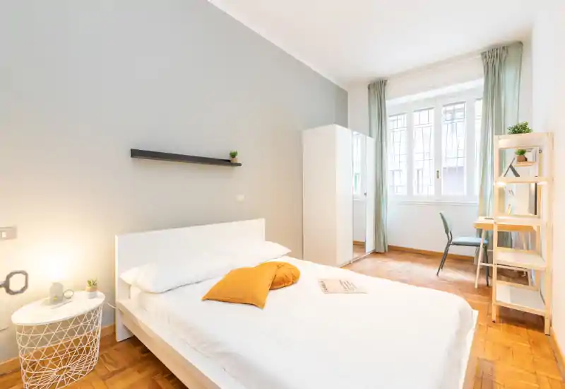 Dormitorio moderno y luminoso con cama blanca, suelos de madera cálida y gran ventana que aporta mucha luz natural. Decoración sencilla y ordenada con cojín y lámpara de mesita.