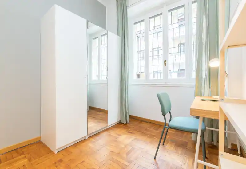 Rincón de dormitorio luminoso y minimalista con suelo de parquet, armario blanco con espejo y pequeño escritorio junto a la ventana — sencillo y bien iluminado, ideal para una persona o estudiante.