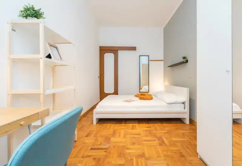 Dormitorio luminoso y moderno con cama blanca, suelo de parquet y estantería minimalista; bien iluminado y amueblado de forma sencilla para una estancia tranquila.