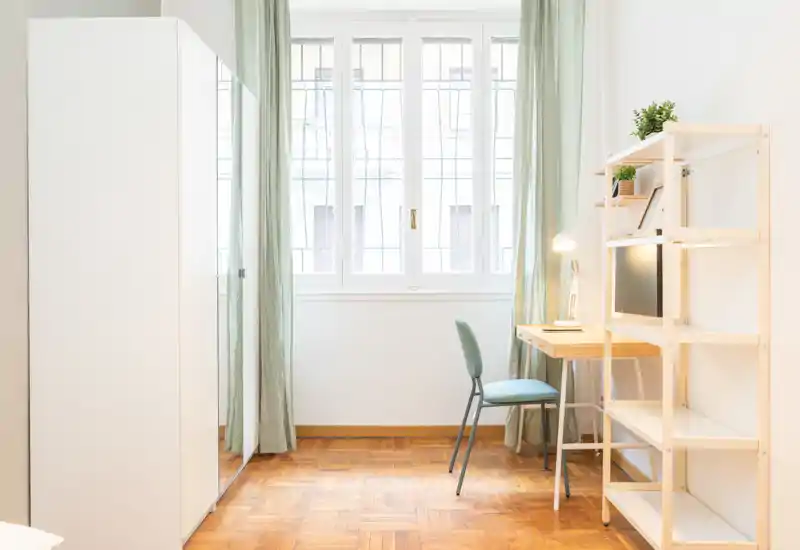 Angolo di camera da letto luminoso e minimale con armadio, scrivania e scaffalatura. La luce naturale entra dalle ampie finestre creando un'area tranquilla per lavoro e riposo.