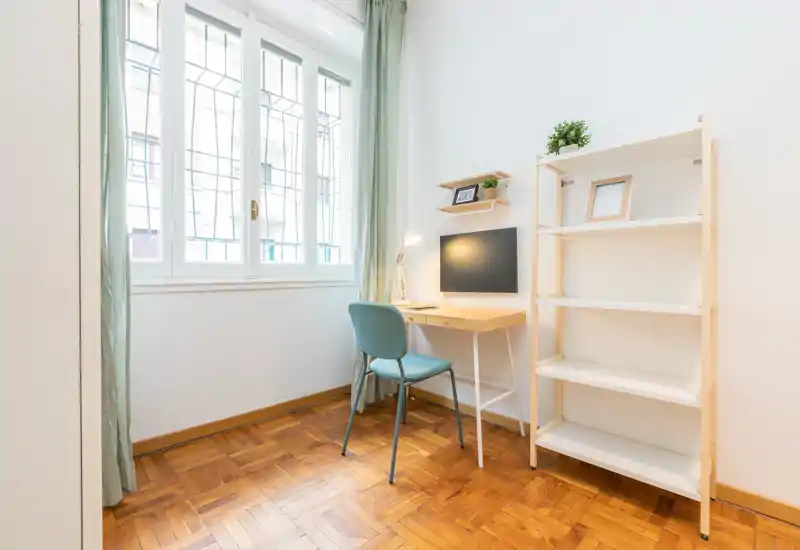 Angolo camera compatto e luminoso con una piccola scrivania vicino a una grande finestra, pavimento in parquet e scaffalatura aperta — perfetto come angolo studio nella zona notte.