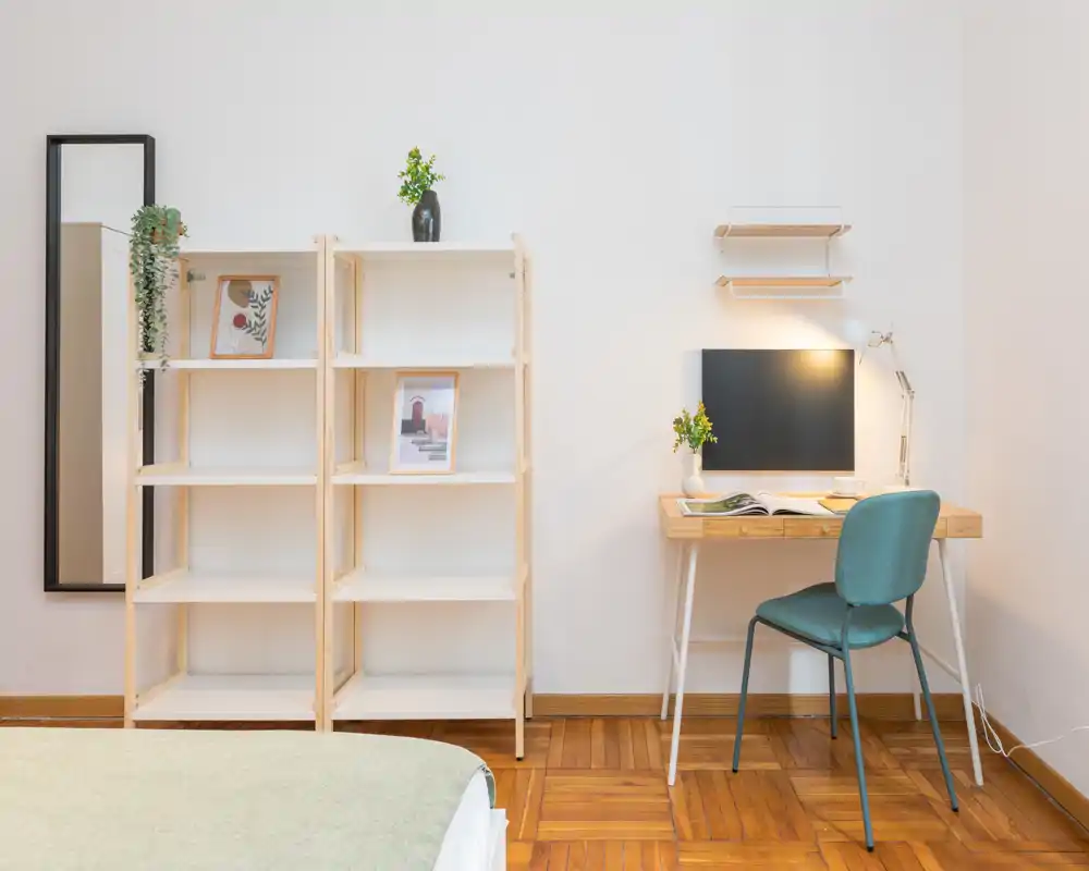Bright, minimalist bedroom corner with a partially visible bed, open shelving and a small desk with a chair — ideal for single occupancy or a student. The space feels clean and well lit with simple decorative touches.
