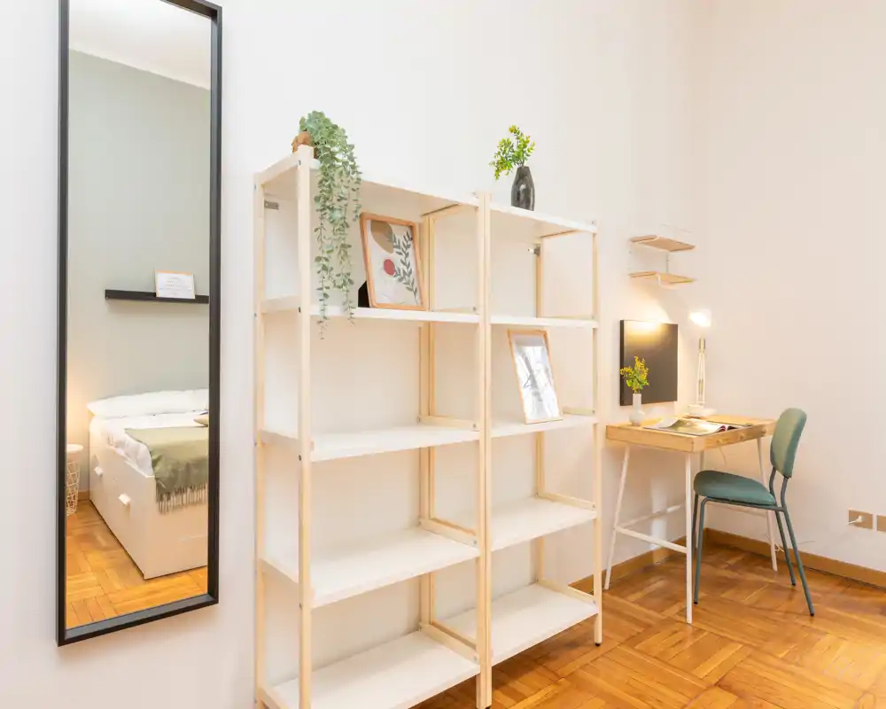 A bright, minimal bedroom corner shown with shelving, a small desk and a tall mirror reflecting the bed. Soft natural light, simple decor and a neat parquet floor create a calm, uncluttered atmosphere ideal for a rental listing.
